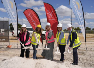 Five people stand near a pile of sand holding shovels. They wear PPE and stand in front of tear-drop banners.