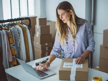 A business owner using her laptop with boxes and clothes nearby